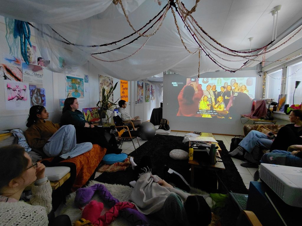 Nuoria kerääntynyt istumaan sohville ruudun ääreen. Ruudusta näkyy video taiteella vaikuttamisesta.